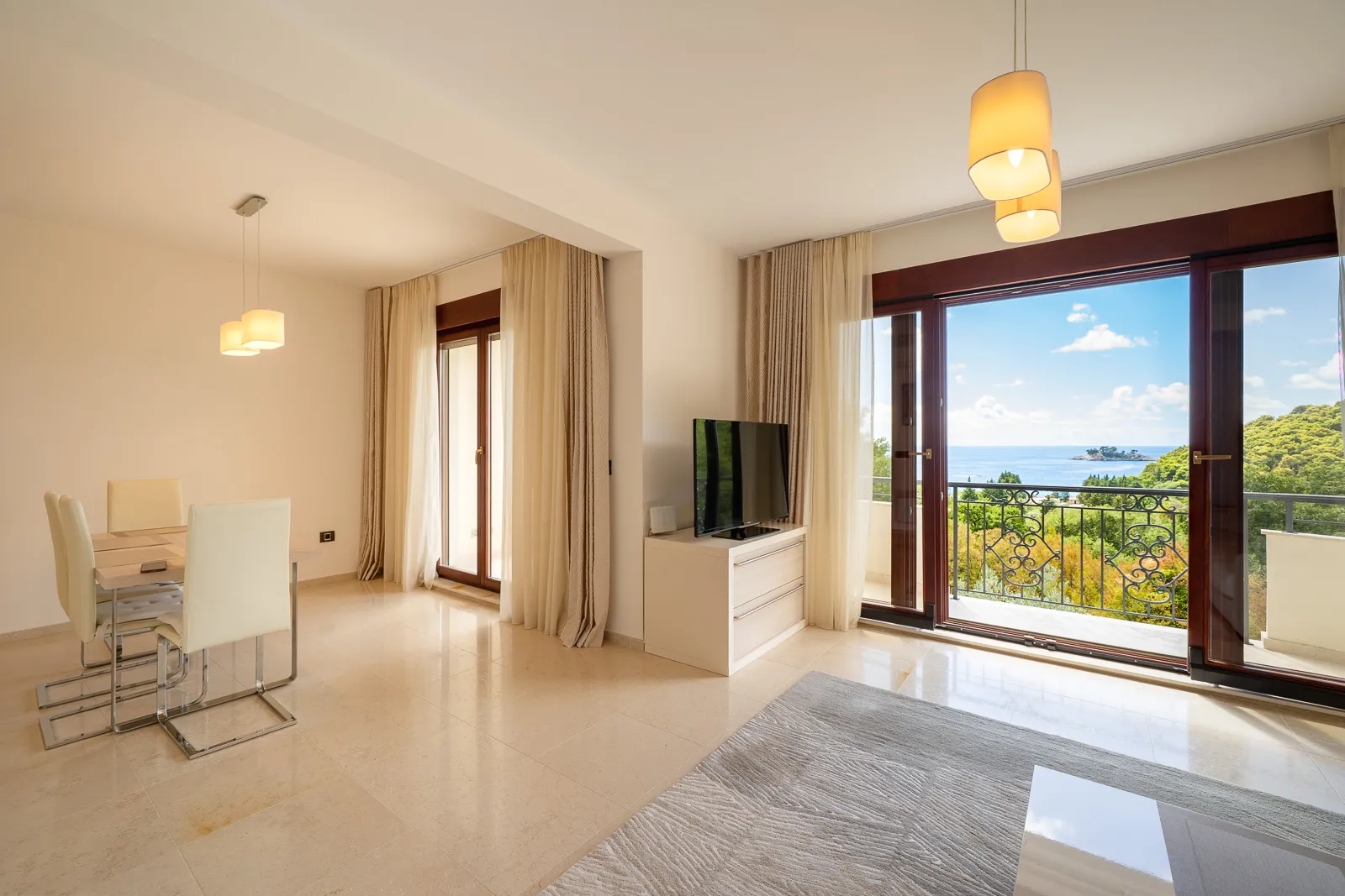 Bright living room with a dining area, TV on a console, and sliding doors opening to a balcony overlooking the sea.