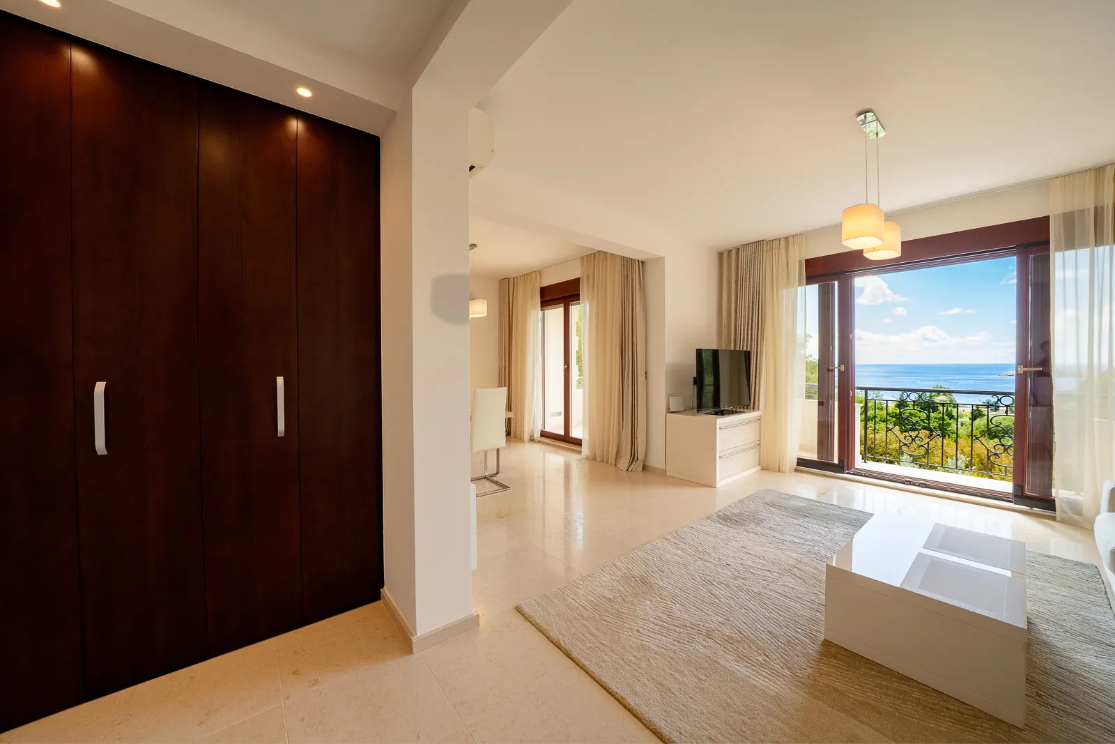 Bright living room with dark wood wardrobe, white coffee table, and TV on a console. Sliding glass door leads to a balcony with sea view.