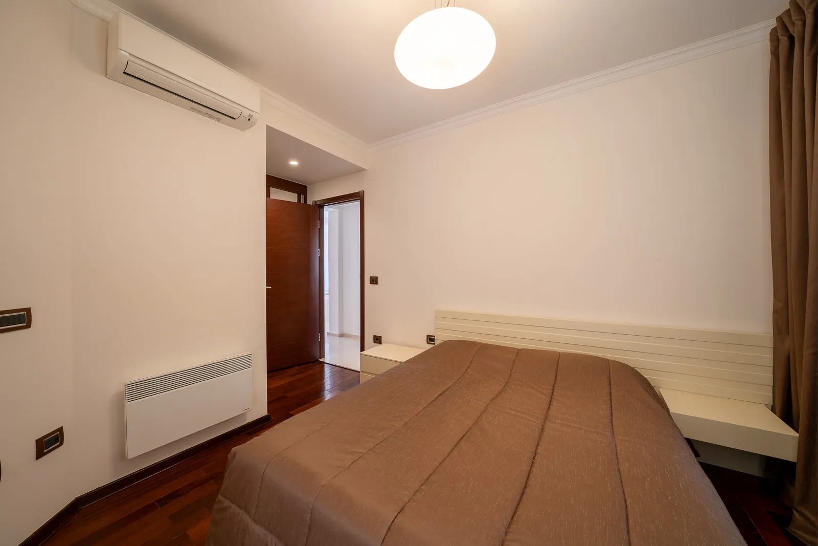 Bedroom featuring a double bed with brown cover, wall AC unit, heater, dark wood flooring, and white walls.
