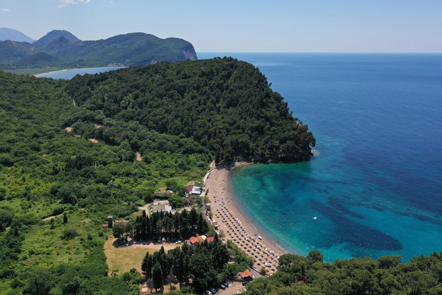Aerial view of Lucice Beach and the Petrovac coastline, Montenegro: azure sea, dense forest, and resort area.