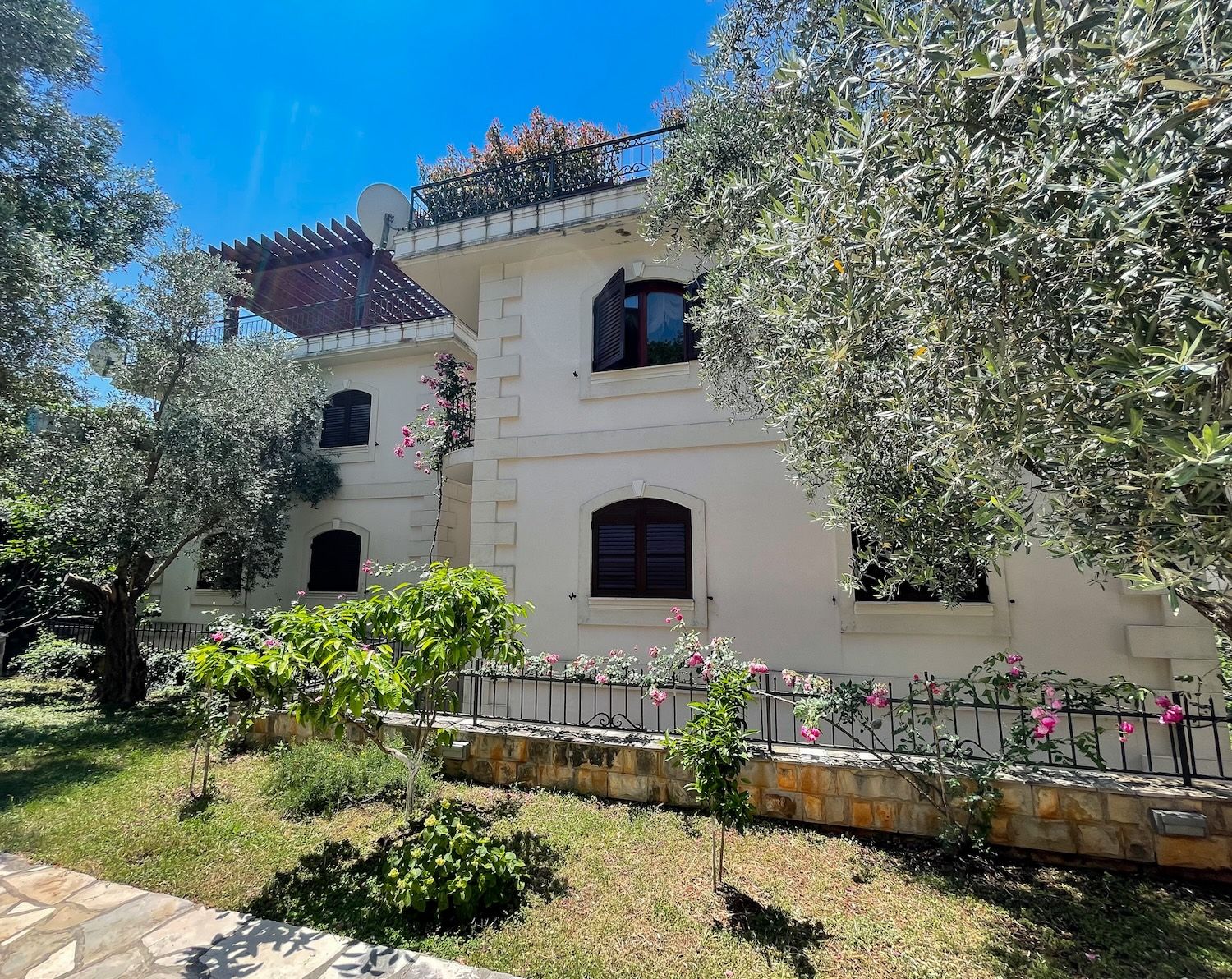 Facade of a three-story villa in Petrovac, Montenegro, with a terrace, olive trees, and a blooming garden on the plot.