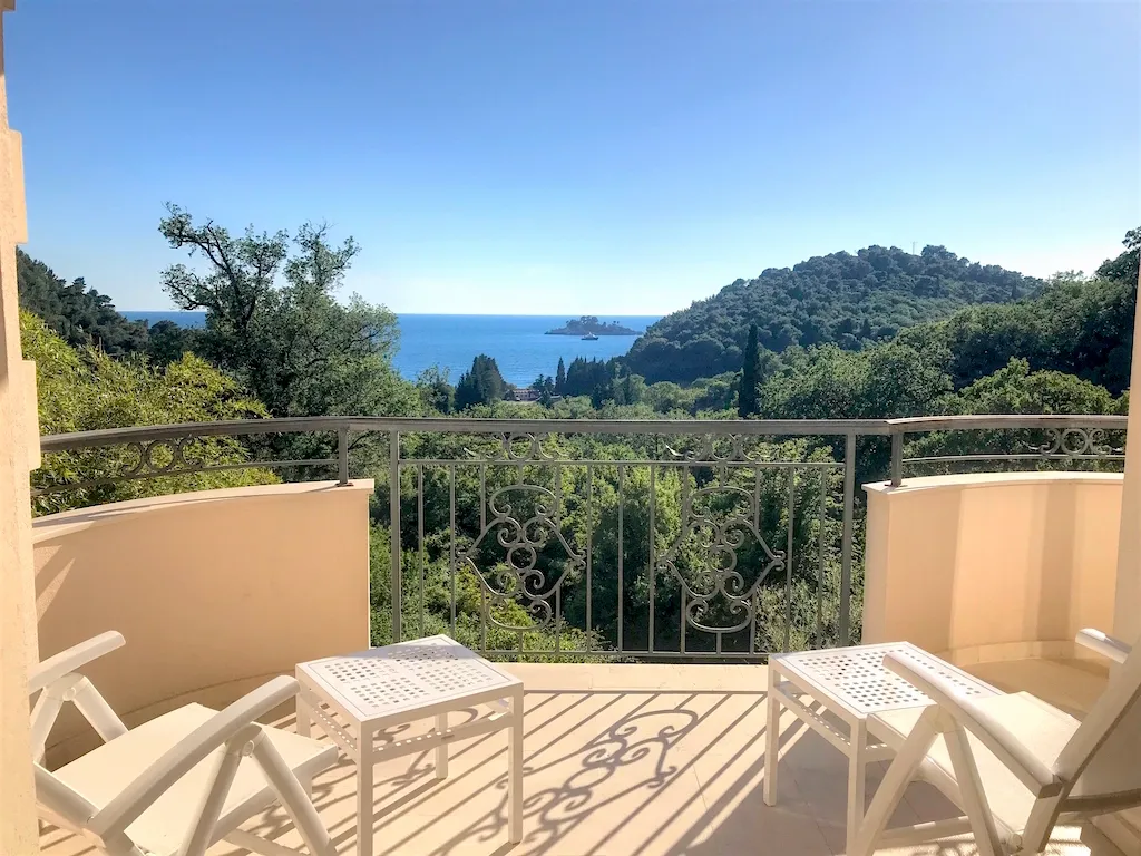 Balcony with white plastic chairs and ornate railing overlooking the sea and a green hill.