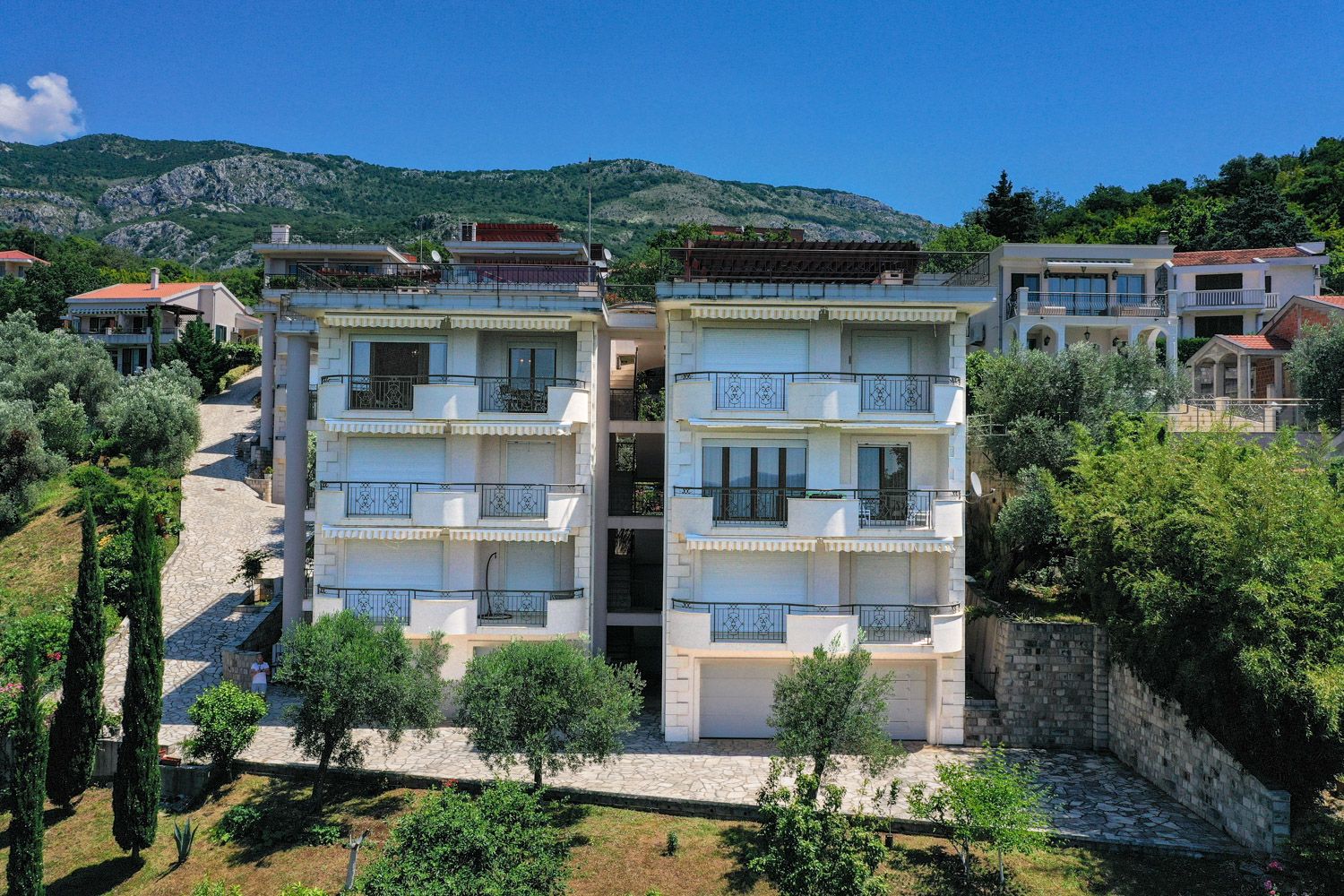 Three-story villa with balconies and terraces in Petrovac, Montenegro: view of the building facade surrounded by greenery and mountains.