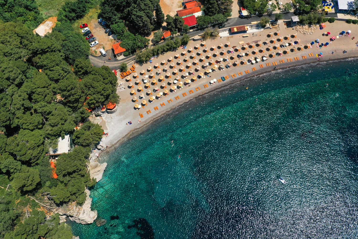 Aerial view of the beach and azure sea in Petrovac, Montenegro: a furnished relaxation area with sun loungers and parking.