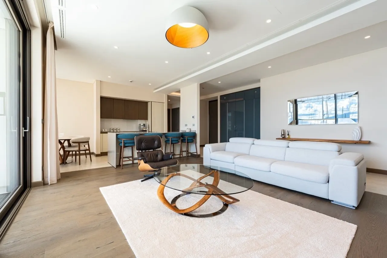 Bright living room with a white sofa, glass coffee table, and open kitchen visible in the background.
