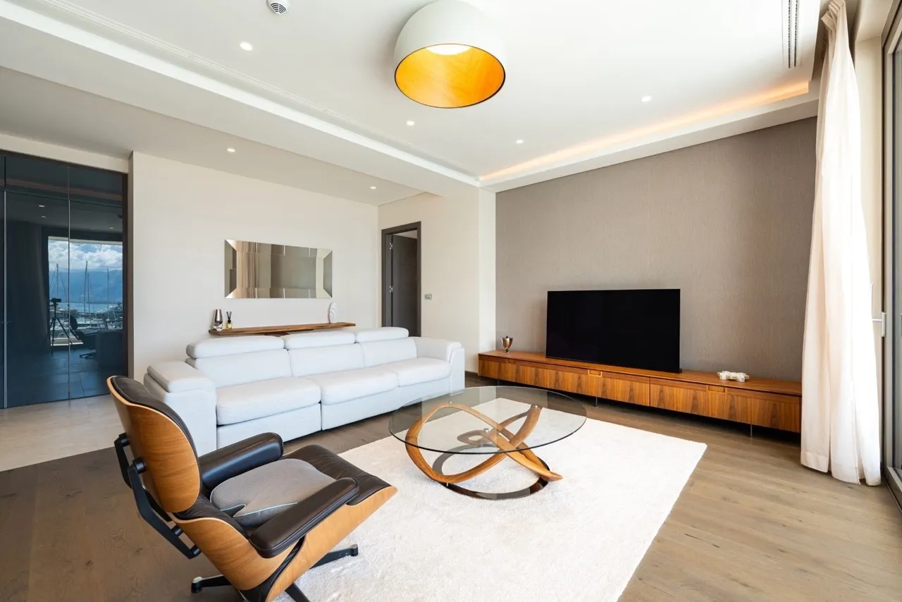 Bright living room with a white sofa, wooden TV unit, glass coffee table, and modern pendant light.