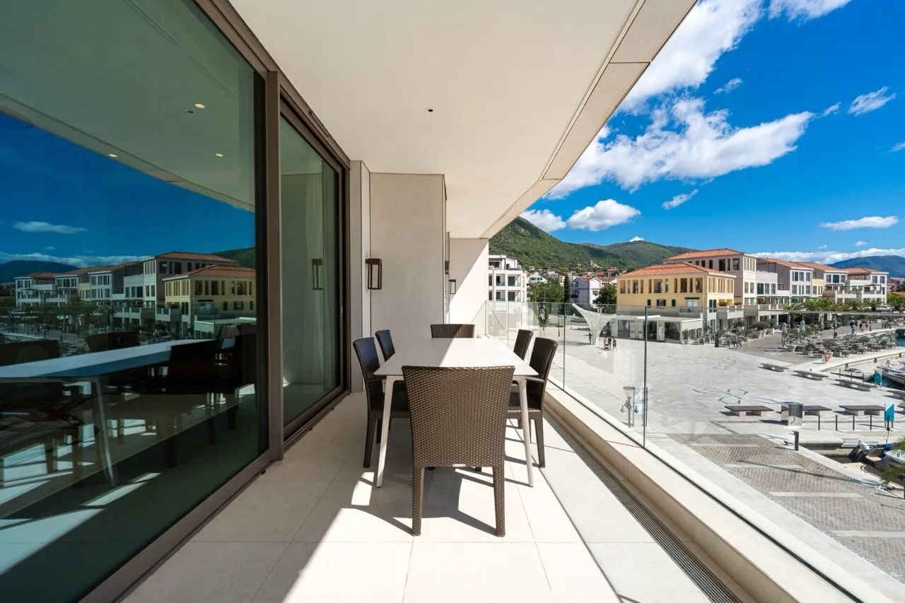 Open balcony with a dining table and wicker chairs overlooking a plaza and mountains through a glass railing.