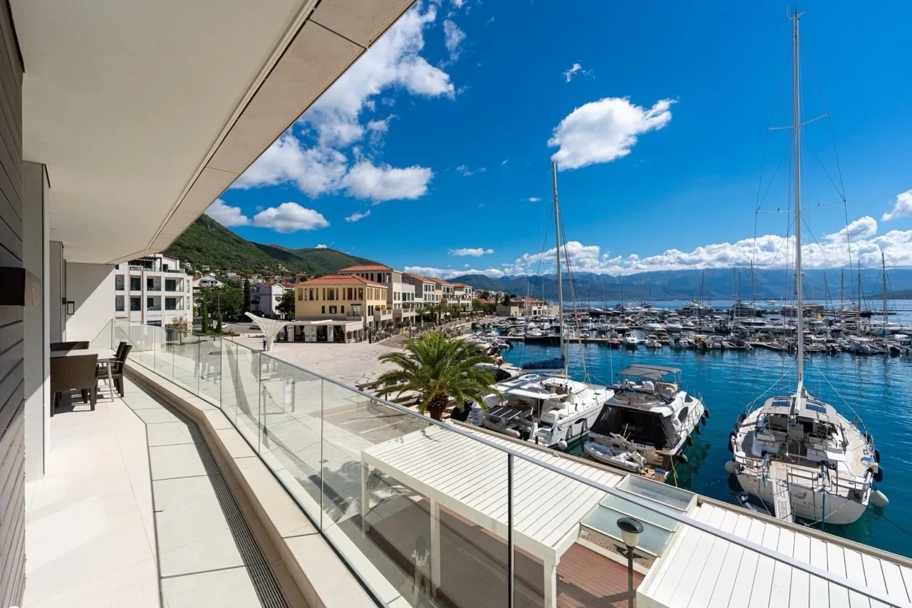 Modern balcony with glass railing overlooking a marina filled with boats, mountains, and sea.