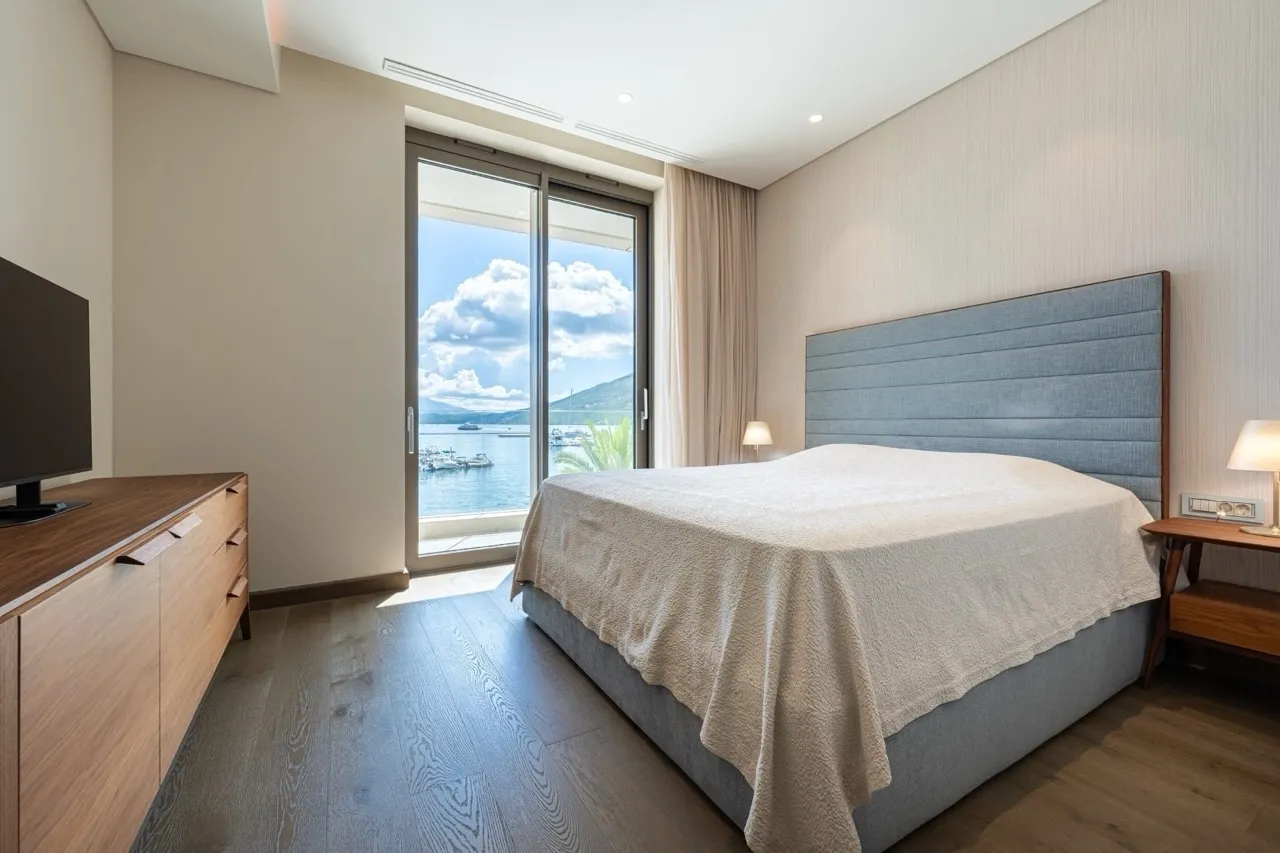 Modern bedroom featuring a large bed with blue headboard, wooden dresser, and sliding glass door to a sea view.