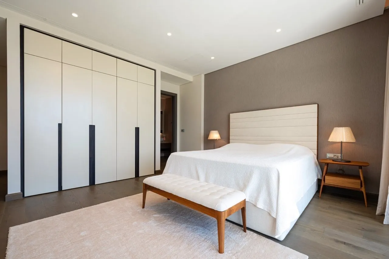 Modern bedroom featuring a large bed, white wardrobe, wooden bench on a rug, and nightstand.
