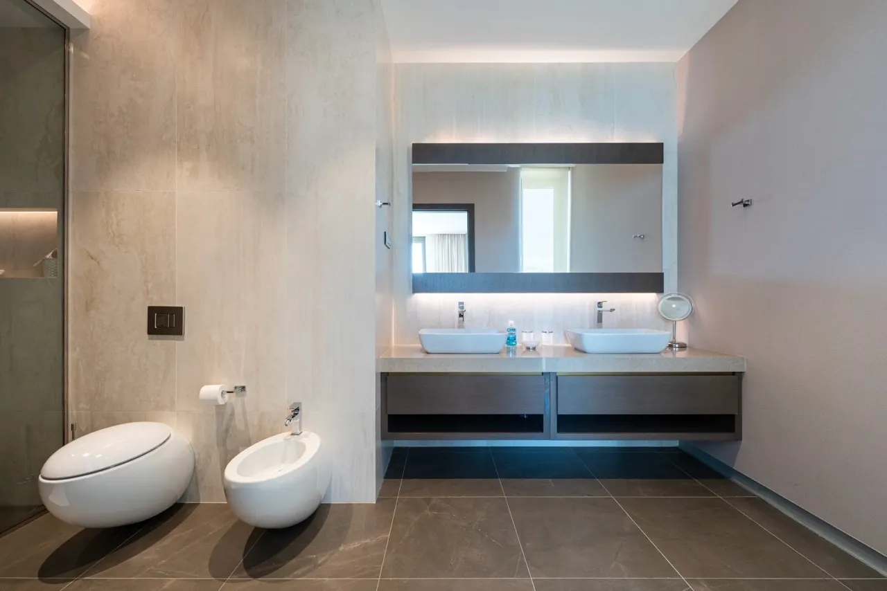 Modern bathroom featuring a wall-hung toilet, bidet, and double vessel sink vanity with large backlit mirror.