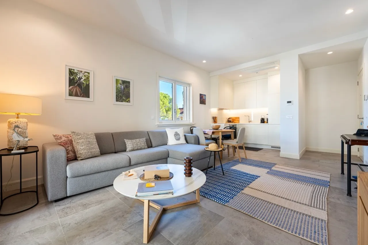 Bright living room with a grey sofa, round table, and blue rug. A white kitchen and dining area are visible in the background.