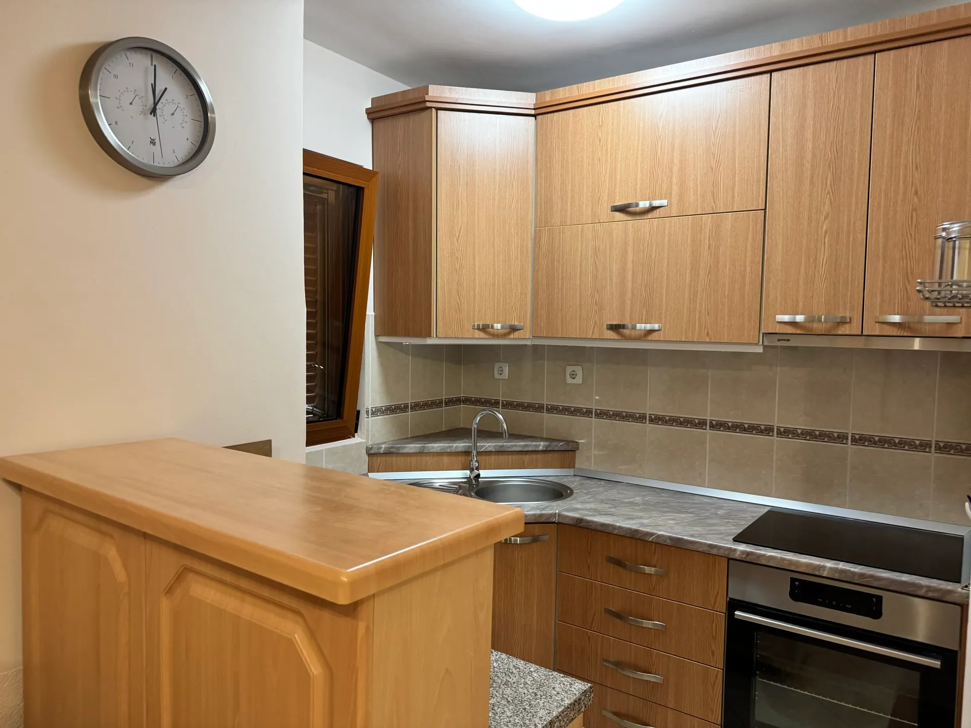 Kitchen with light wood cabinets, grey countertop, sink, and built-in oven under a black cooktop.