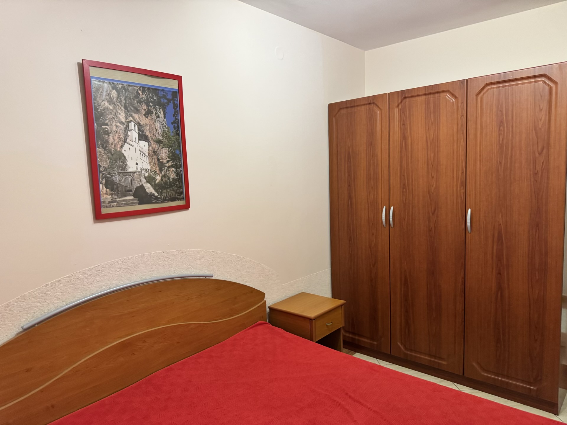 Bedroom in an apartment in Budva, Montenegro: a double bed, a wooden wardrobe, and a painting of Ostrog Monastery.