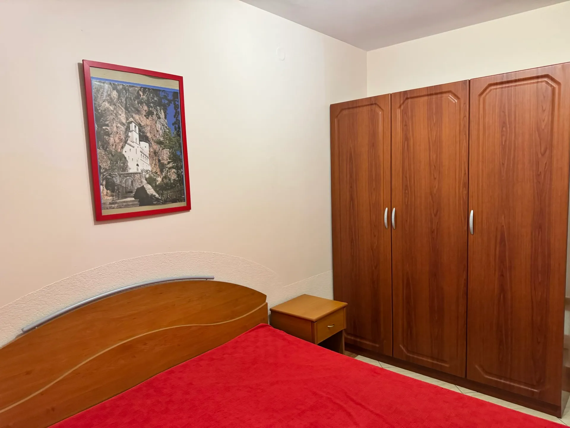 Bedroom featuring a wooden bed with red cover, large wardrobe, small nightstand, and a framed picture on the wall.