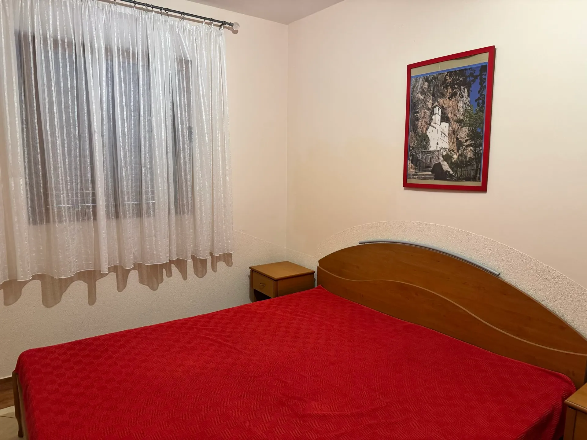 Bedroom with a bed covered in red fabric, wooden headboard, white sheer curtains, and a framed picture.