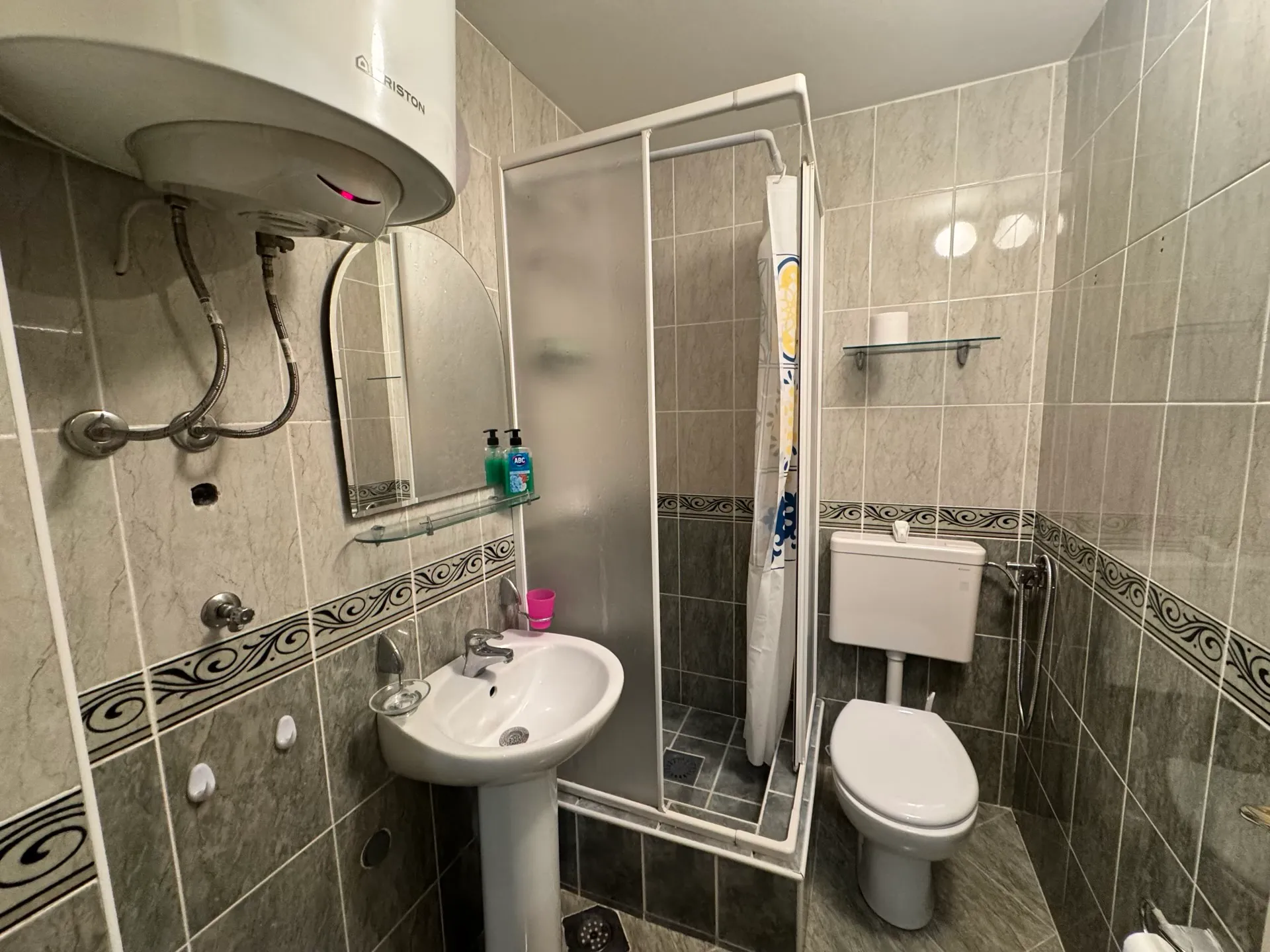 Small bathroom with grey tiles, pedestal sink, shower enclosure, toilet, and wall-mounted water heater.
