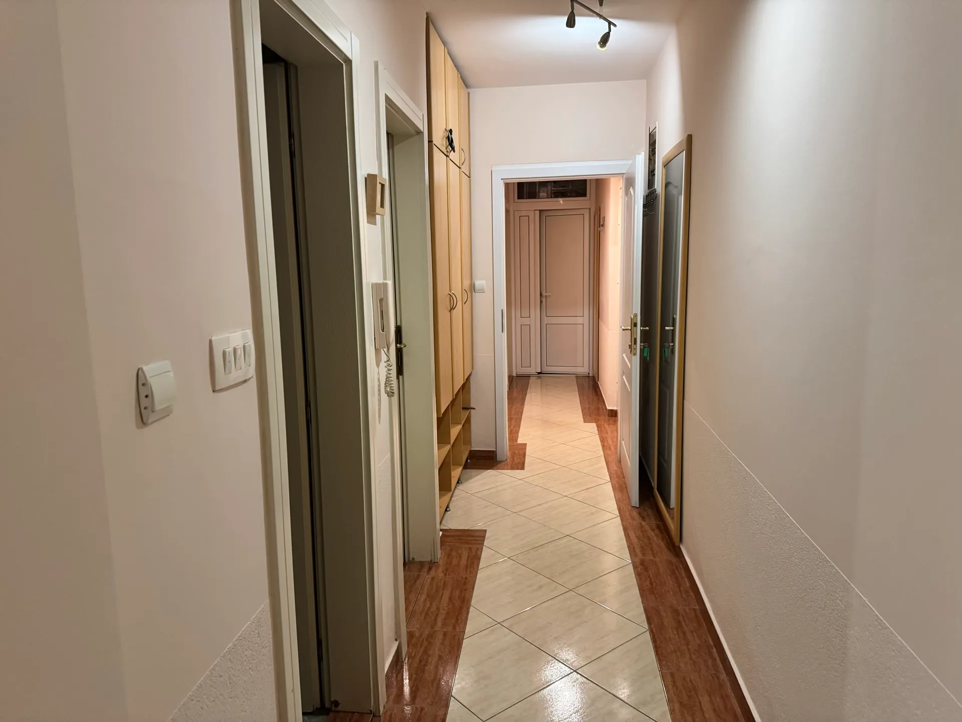 Long hallway with light tiled flooring, tall wooden cabinets on the left, a mirror on the right wall, and multiple doors.