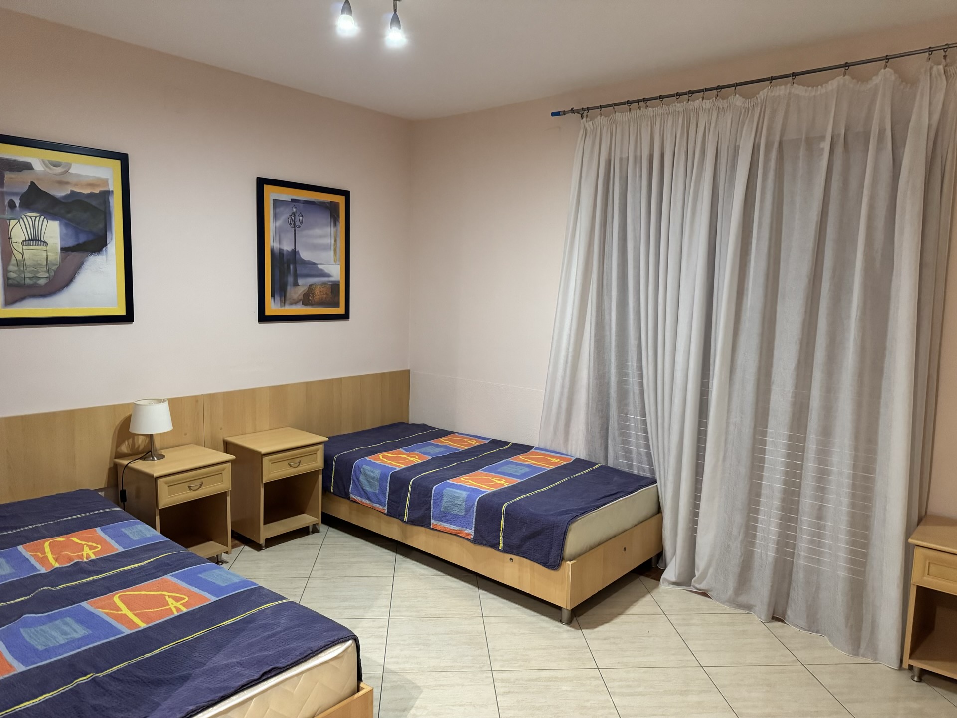 Bedroom with two single beds in an apartment in Budva, Montenegro: light-colored furniture, paintings, and floor-to-ceiling curtains.