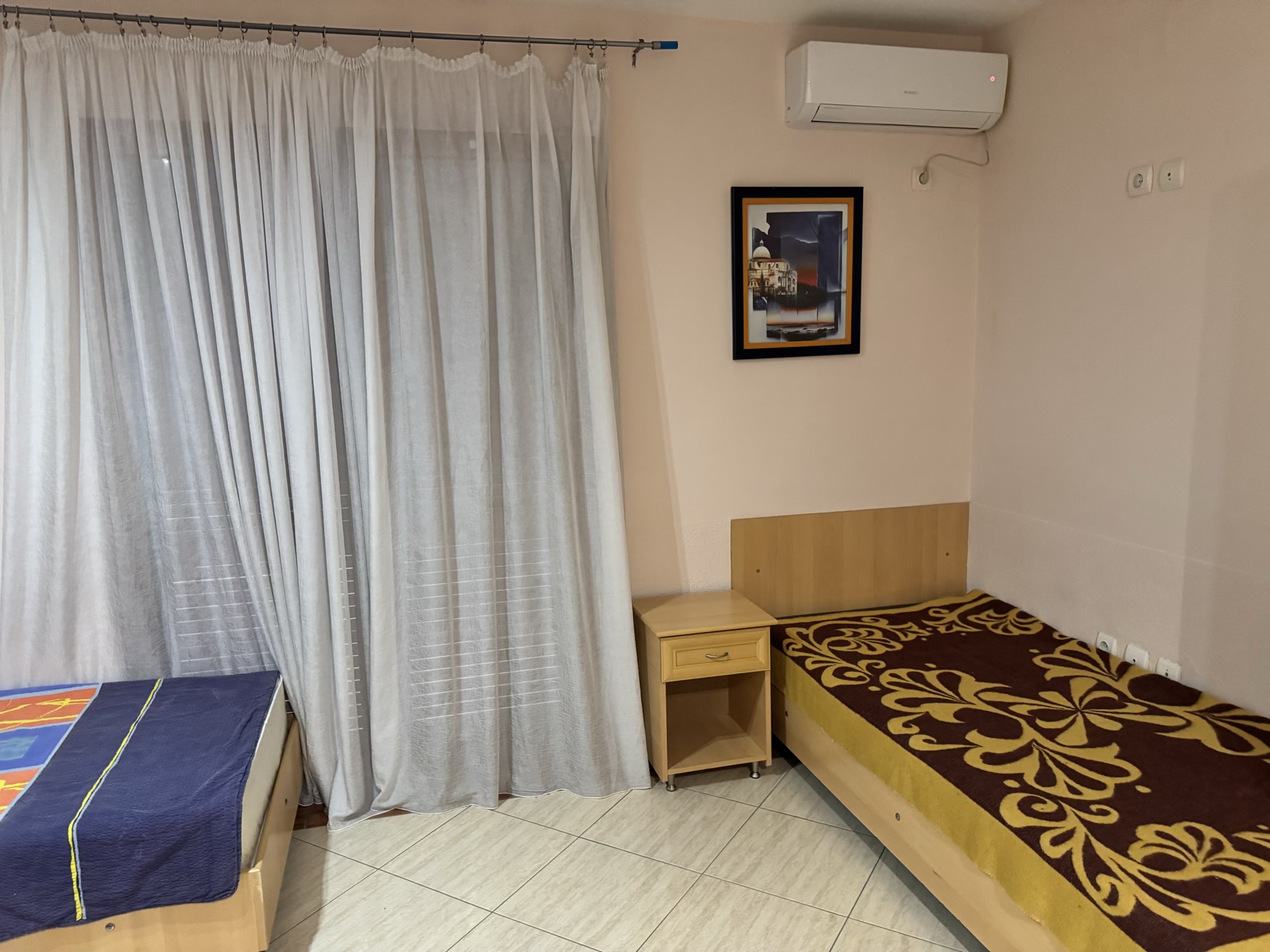Bedroom in an apartment in Budva, Montenegro: two beds, a bedside table, light walls, air conditioner, and panoramic curtains.