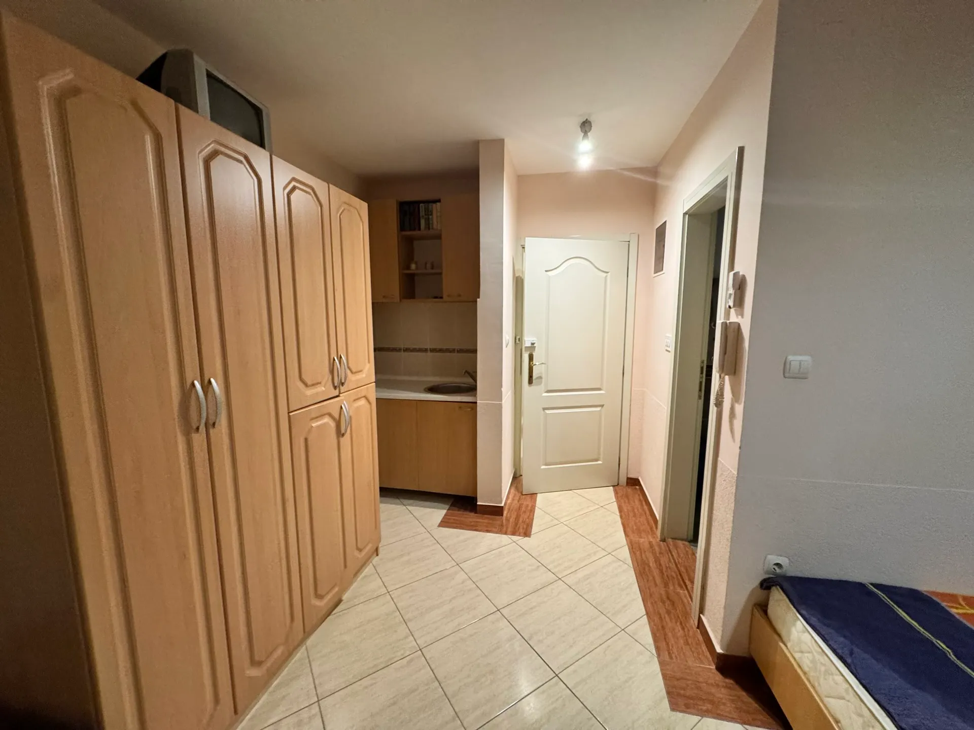 Hallway with wardrobe, partial kitchen view, and bed edge. White door and tiled floor visible.