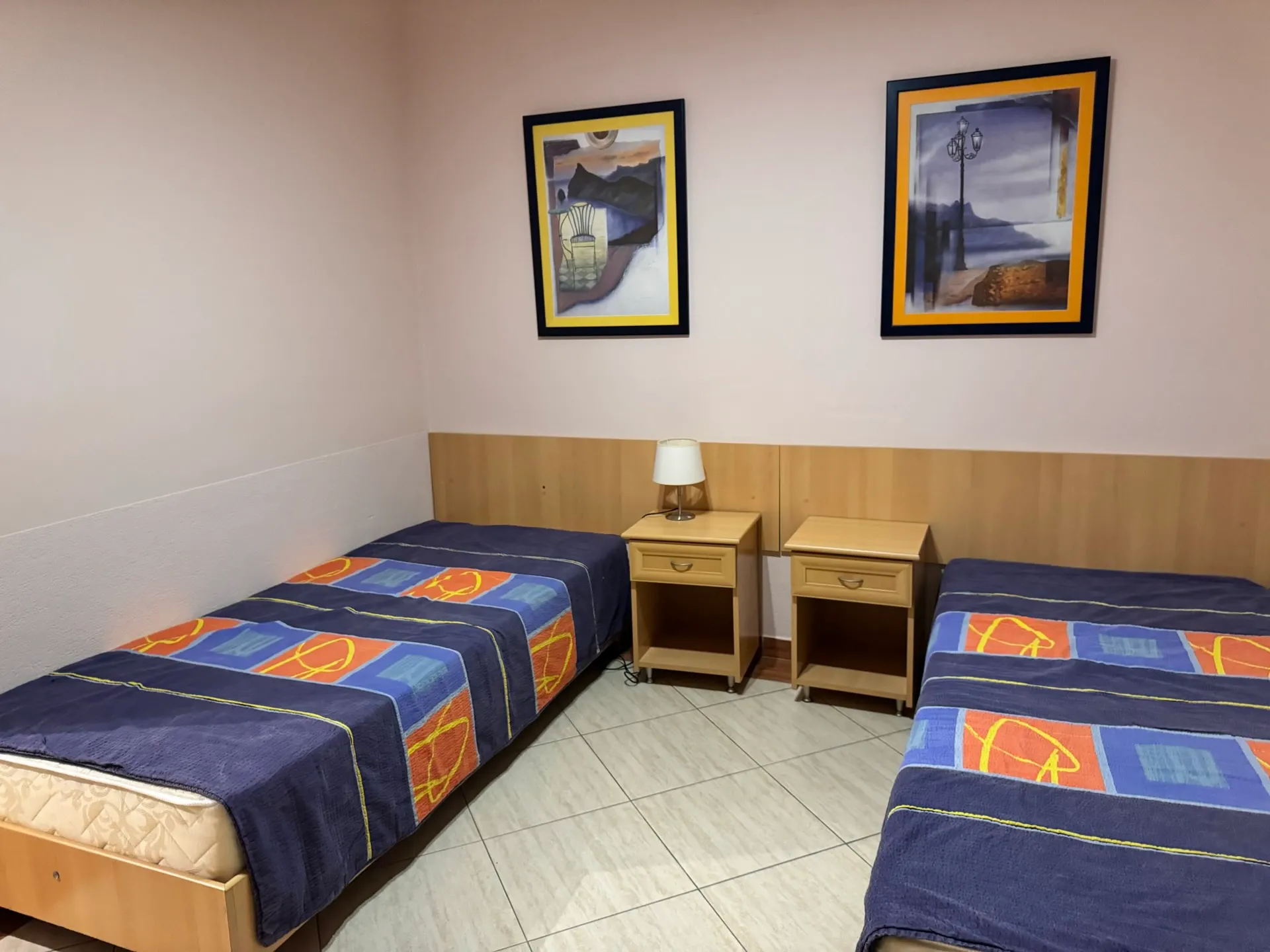 Two single beds with blue covers, wooden nightstands with a lamp, and framed art on the wall.