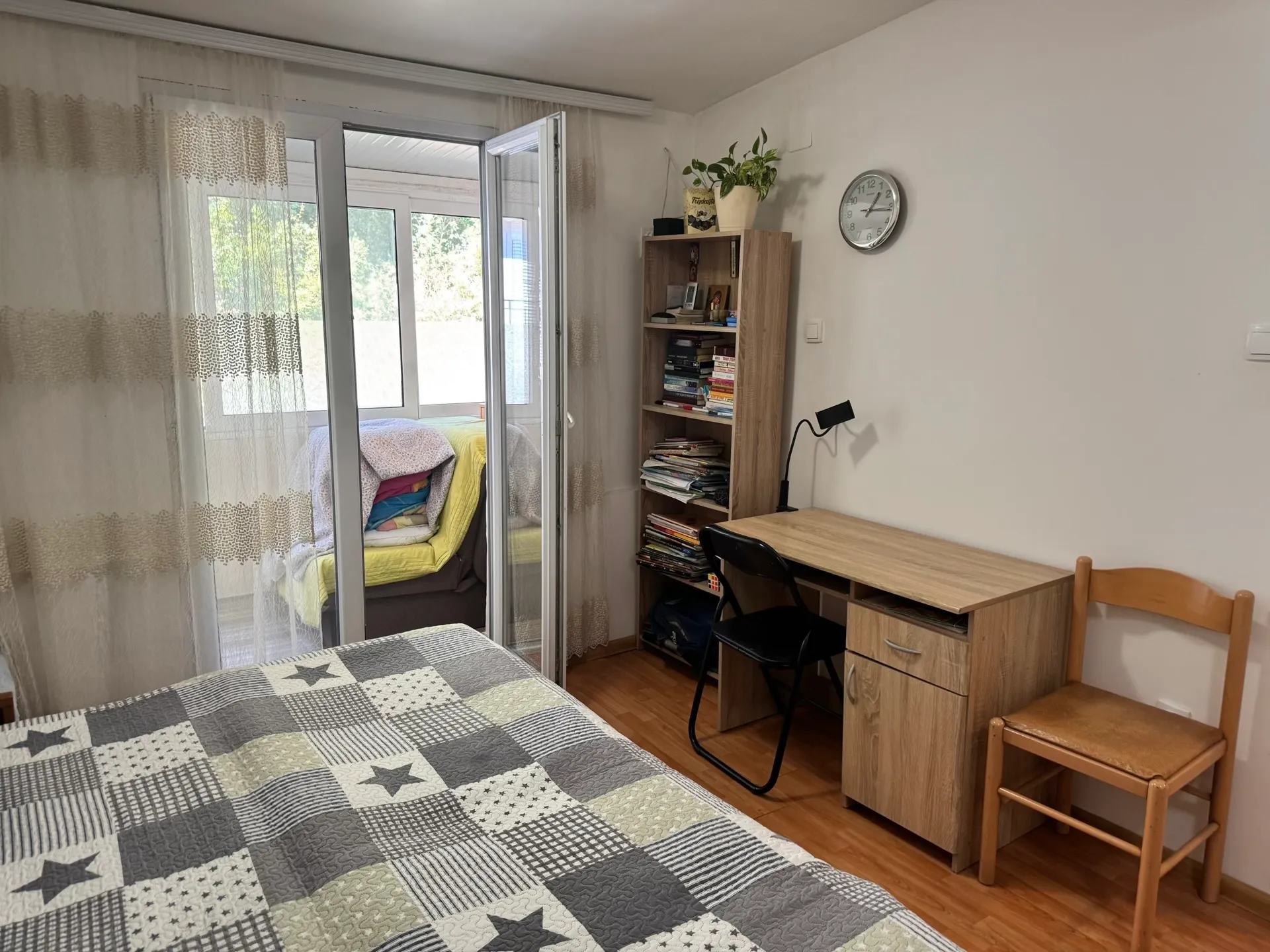 Bedroom featuring a star-patterned quilt, wooden desk, bookshelf filled with books, and an open door to a balcony.
