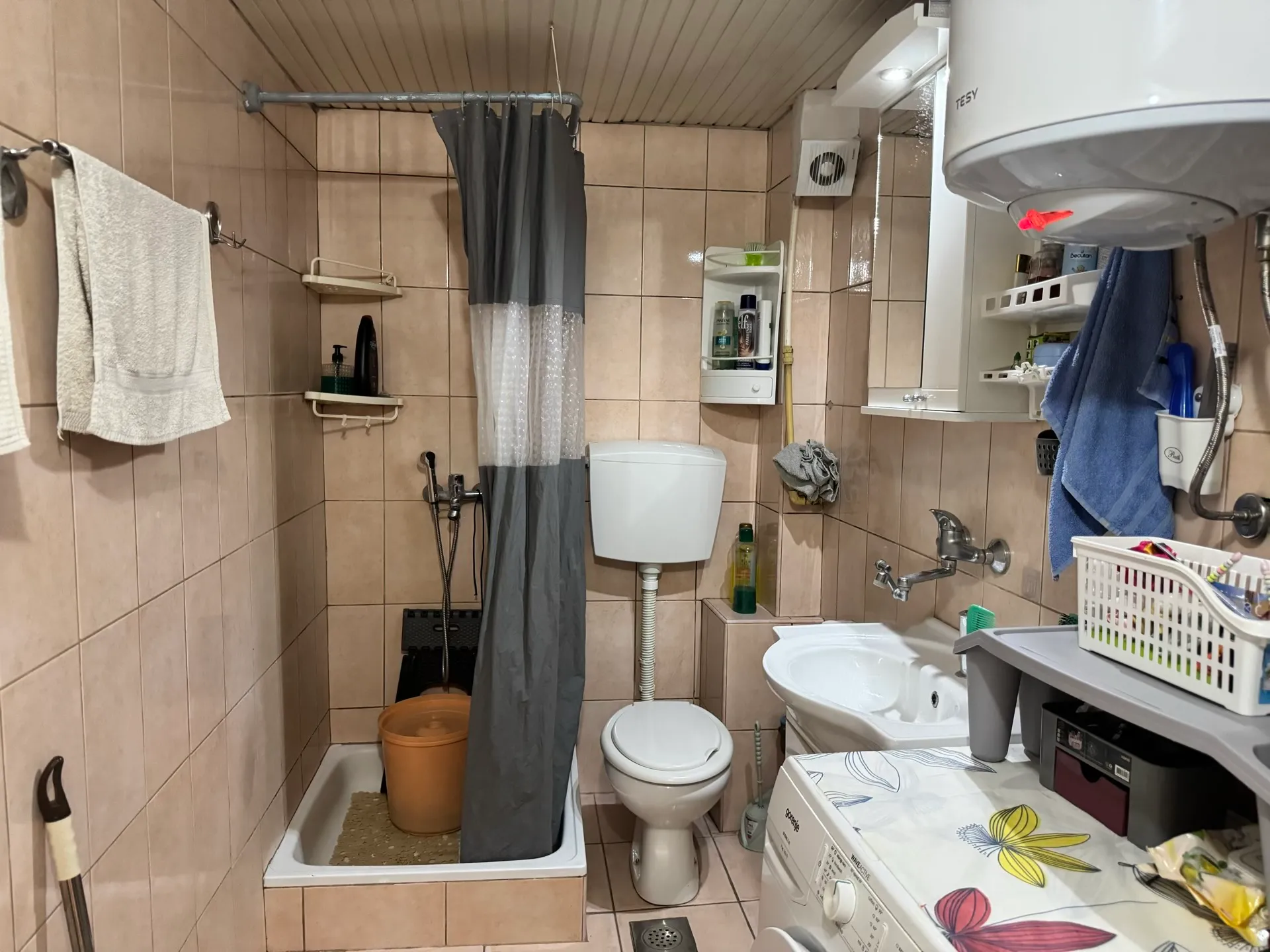 Small bathroom with shower, toilet, sink and washing machine on beige tiles, featuring a grey curtain and wall-mounted water heater.