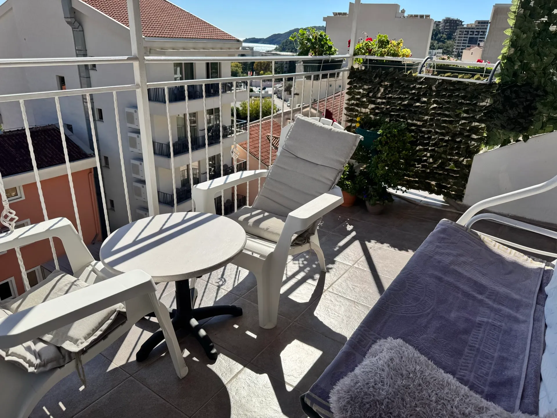 A sunny balcony with white plastic furniture, a small table, and a view of the city and distant sea.