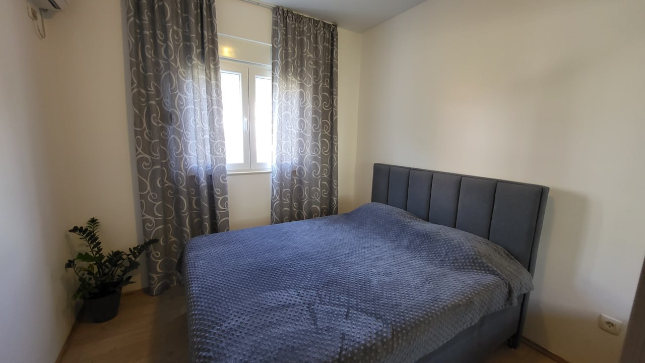 Cozy bedroom with a double bed in an apartment in Bečići, Montenegro: gray textiles, light walls, and a live plant.