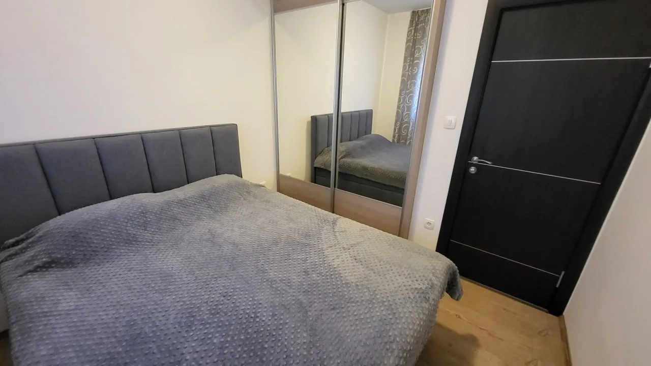 Bedroom featuring a double bed with a grey cover, a mirrored sliding wardrobe, and a black door on light wooden flooring.