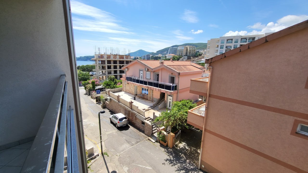 View from the apartment balcony in Bečići, Montenegro: a panorama of neighboring buildings, the street, and the sea coast in the distance.