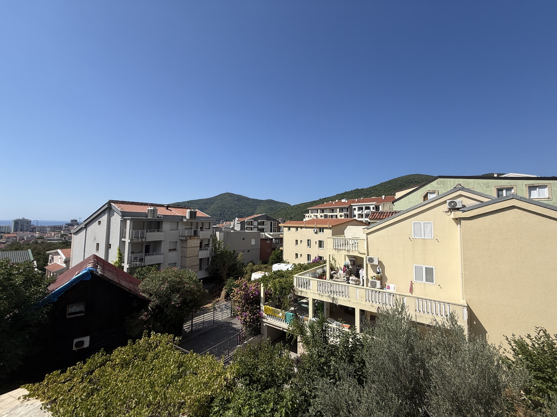 View of a residential area and mountains from an apartment in Budva, Montenegro: low-rise buildings, terraces, and a green garden.