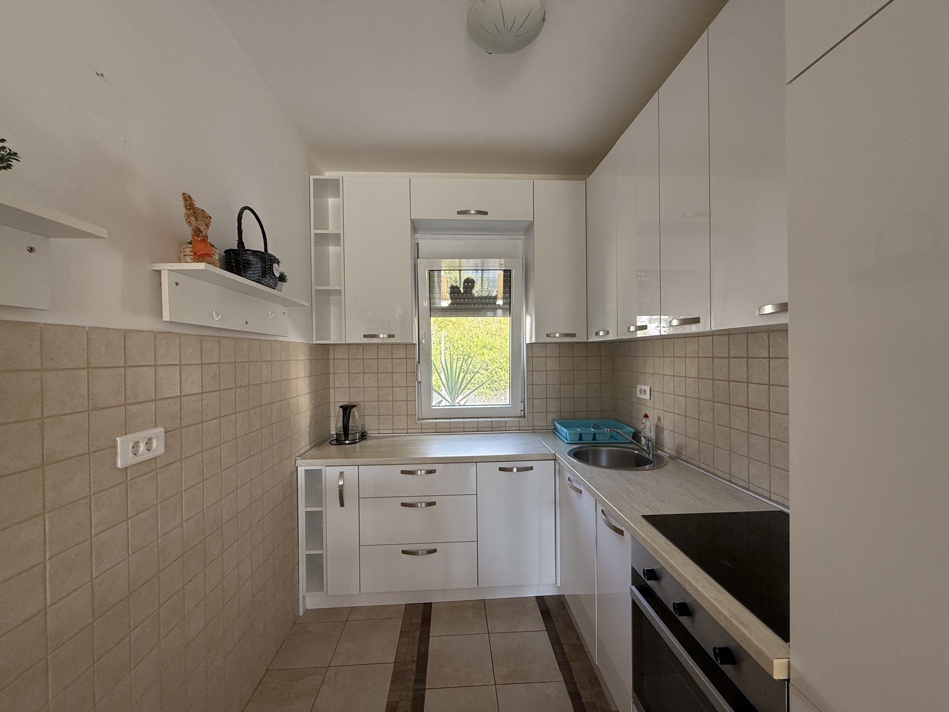 White modern kitchen in an apartment in Budva, Montenegro: built-in appliances, glossy fronts, and a window view of the garden.
