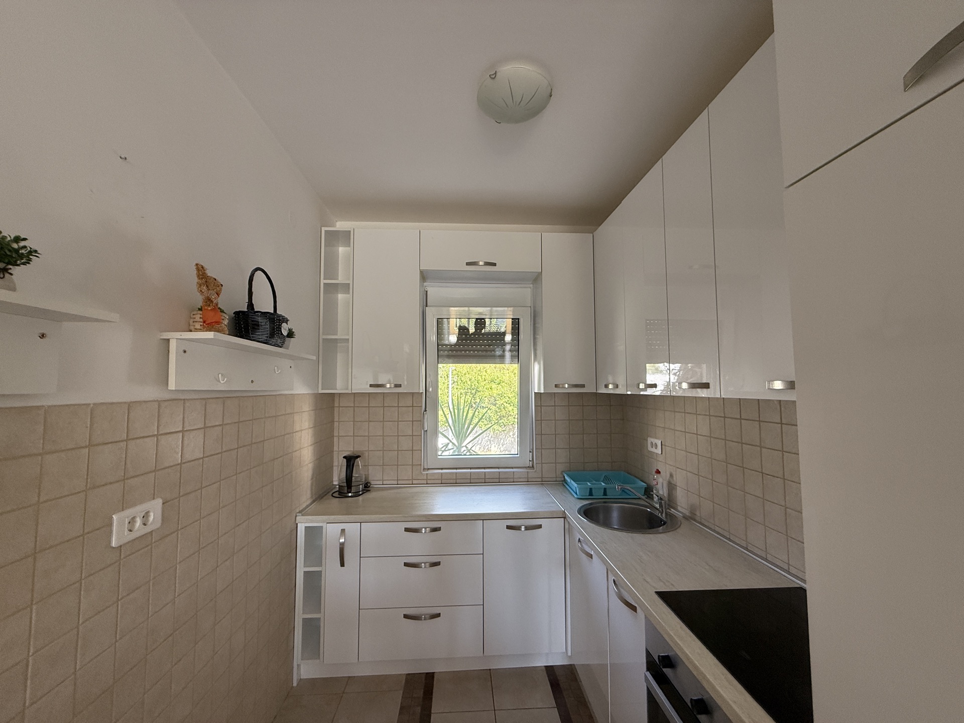 Bright kitchen with white cabinetry in an apartment in Budva, Montenegro: built-in appliances, tile, and a view of greenery from the window.