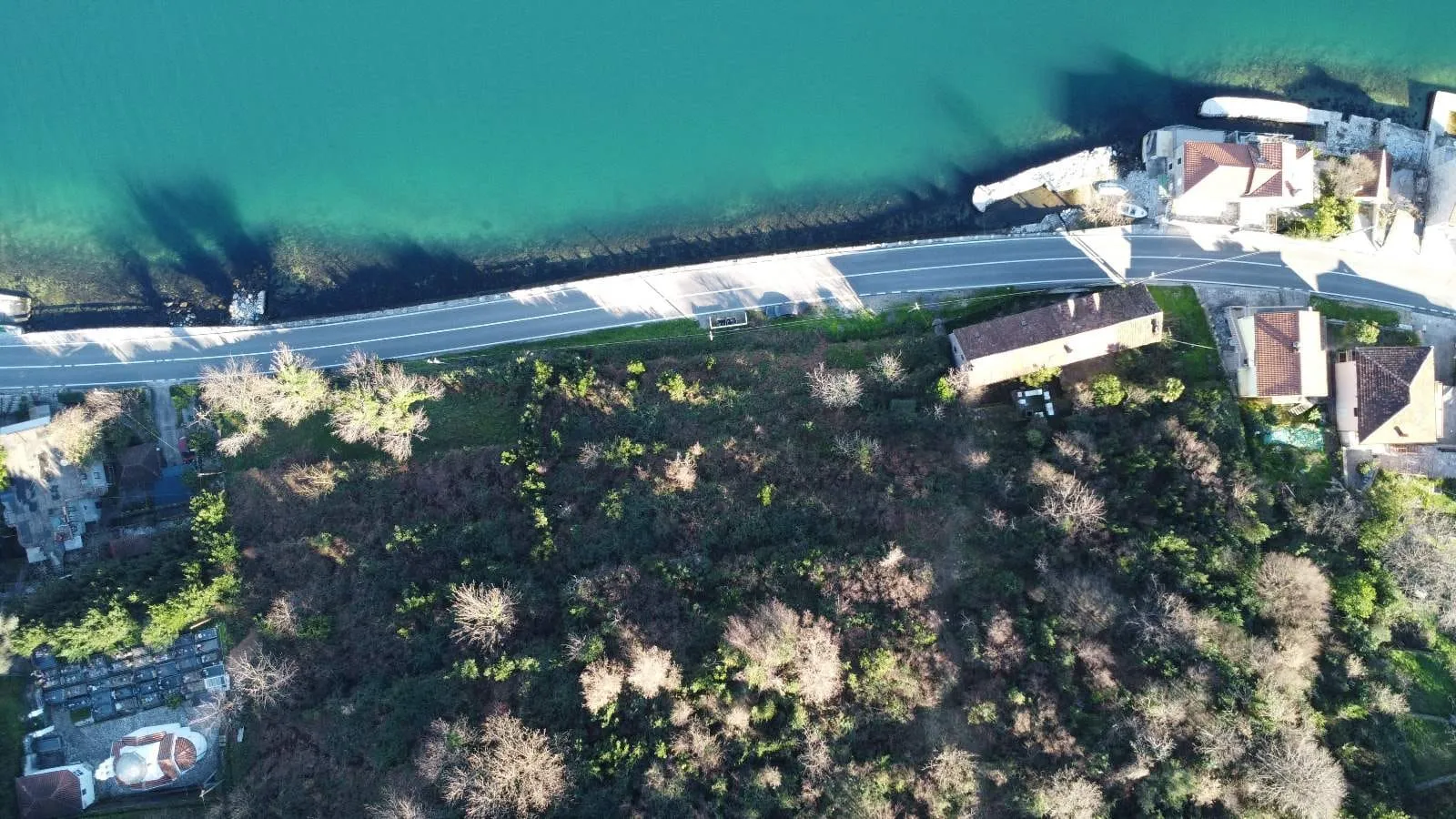 Aerial view of a coastline with turquoise water, a road running alongside, dense forest, houses with red roofs, and a stone embankment.