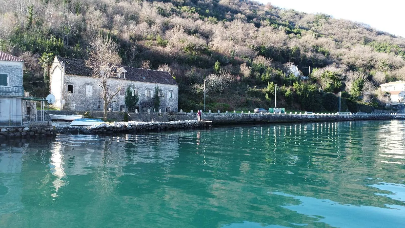Old stone house by turquoise water with a stone quay and forested hill in the background.