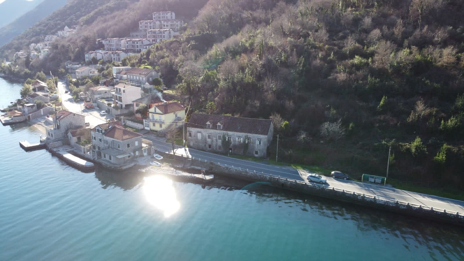Aerial view of a stone house by the sea in the village of Kostanica, Montenegro: coastline, road, and mountain landscape.