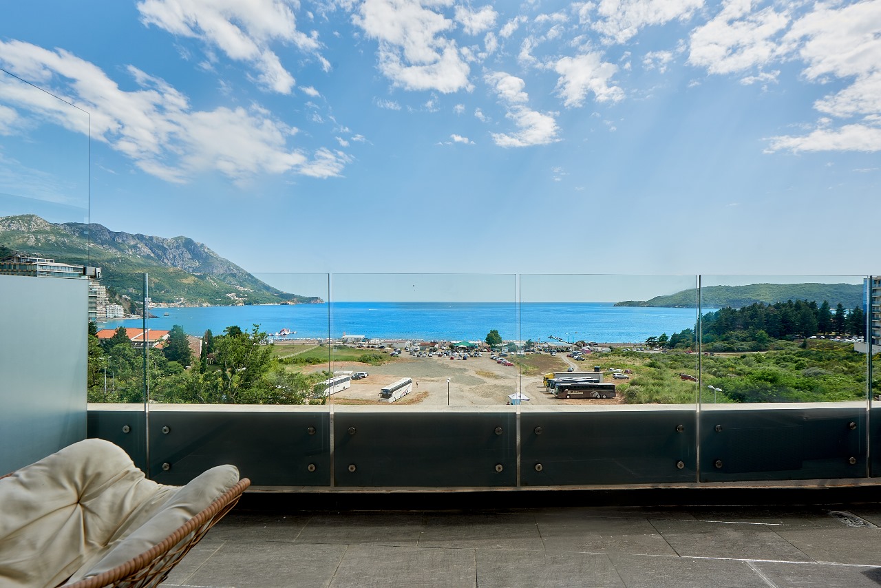 View from the apartment balcony in Bečići, Montenegro: panoramic sea, mountain, and beach views, glass railing, wicker chair.