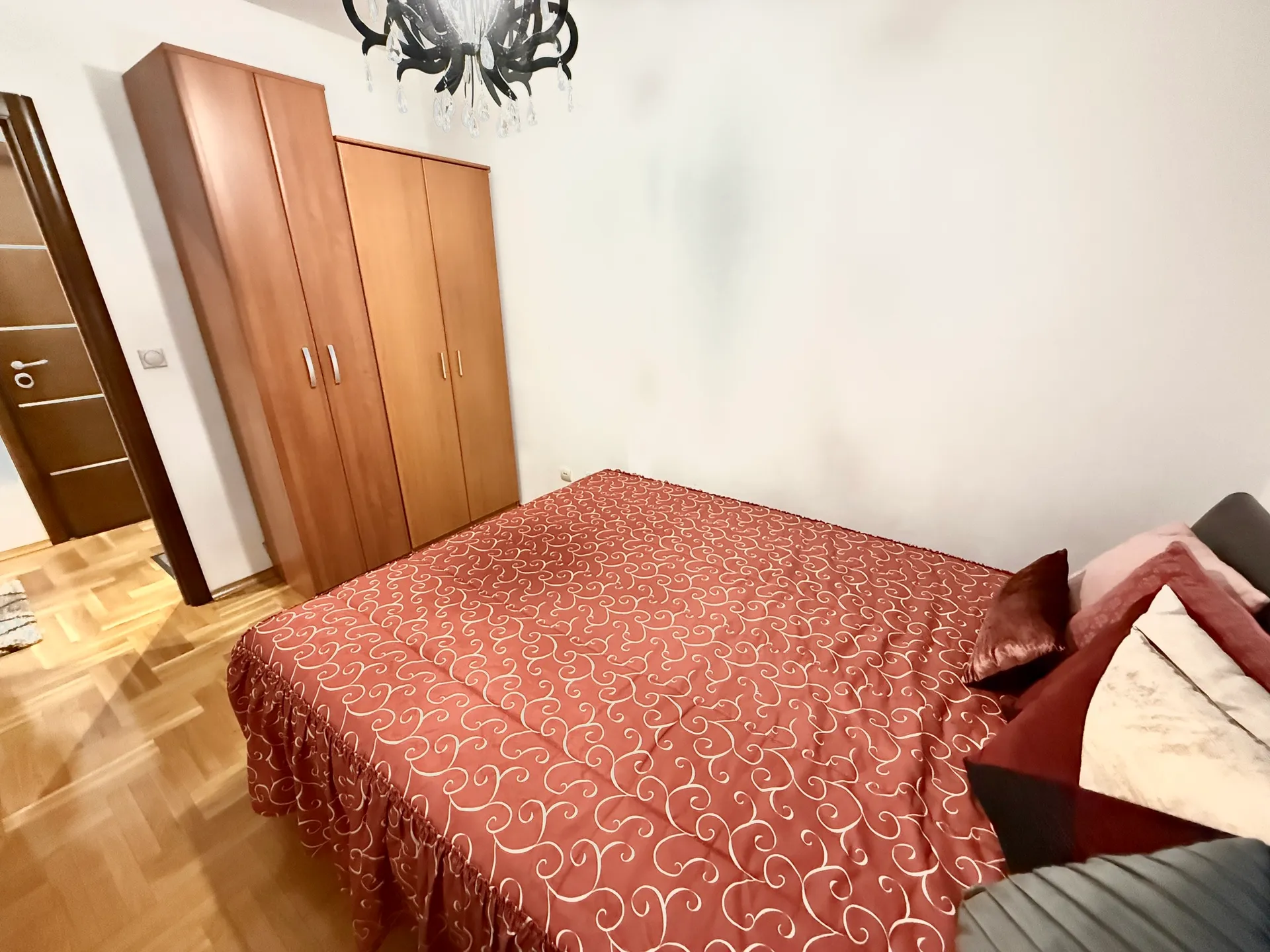 Bedroom featuring a bed with red patterned cover, wooden wardrobes, and a chandelier over parquet flooring.