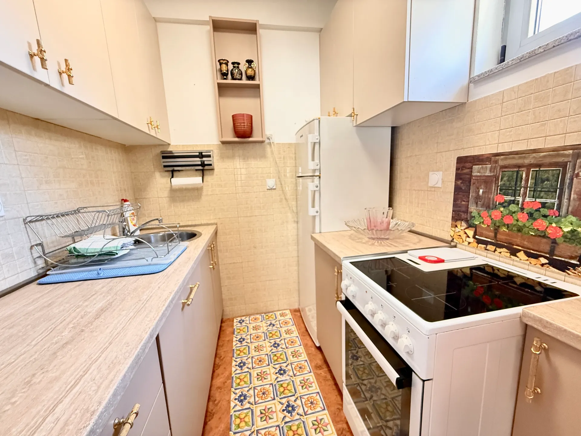 Narrow kitchen with beige cabinets, white wall tiles, an electric stove, and a colorful geometric patterned runner rug on the floor.