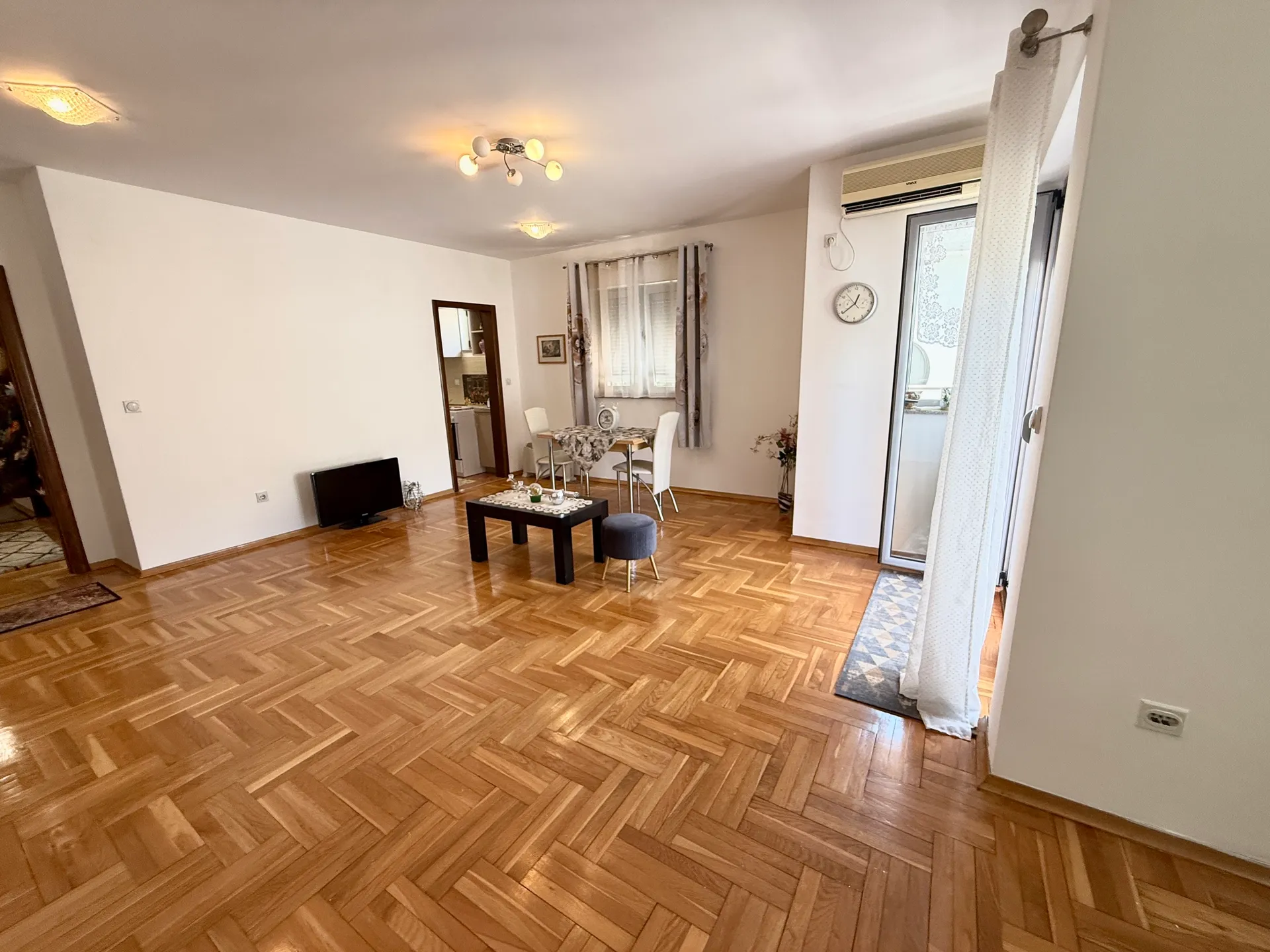 Living room with herringbone parquet floor, black coffee table, TV on the floor, and dining area in the background.
