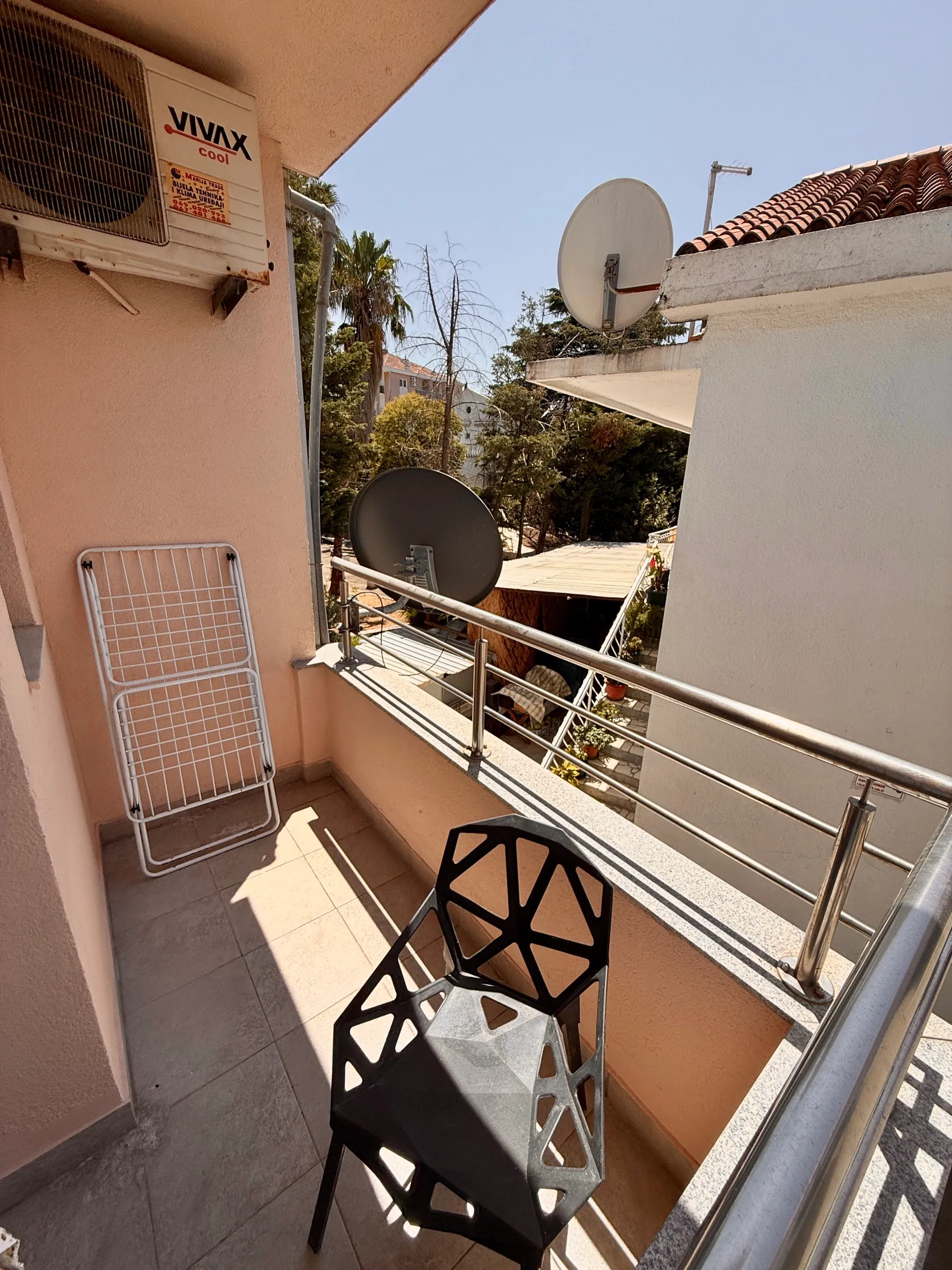 Balcony with a black geometric chair, white drying rack, and wall-mounted air conditioner under bright sunlight.