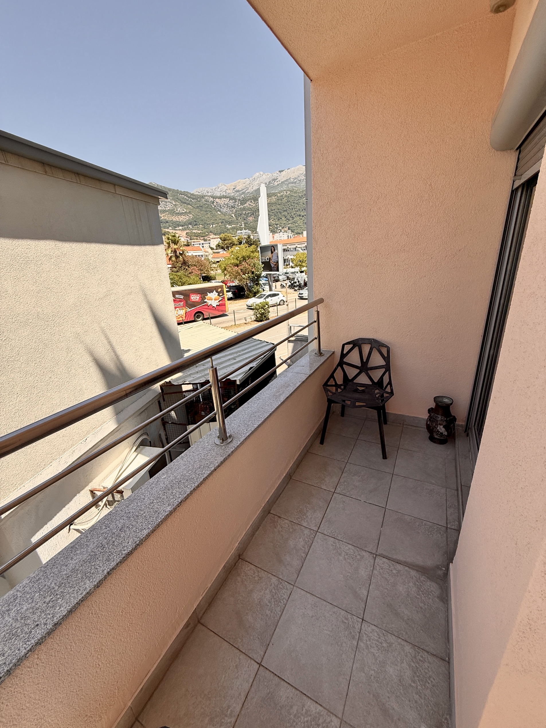 Apartment balcony in Budva, Montenegro: tiled floor, modern chair, view of the mountains and city infrastructure.