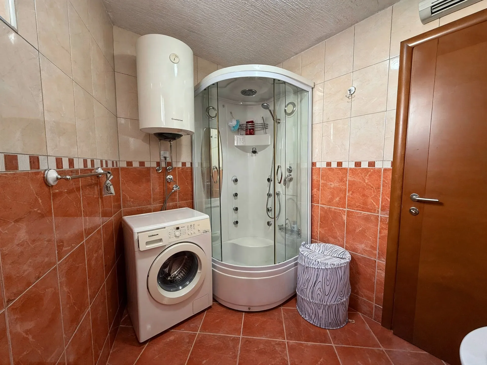 Bathroom featuring a corner shower cabin, washing machine, and wall-mounted water heater amidst orange tiled walls.