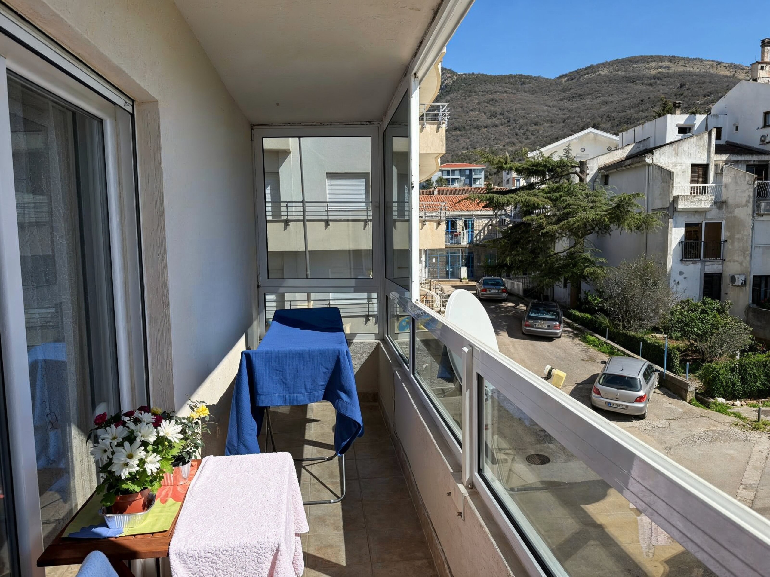 Glazed balcony in an apartment in Bečići, Montenegro, with a view of the street, mountains, and residential buildings.
