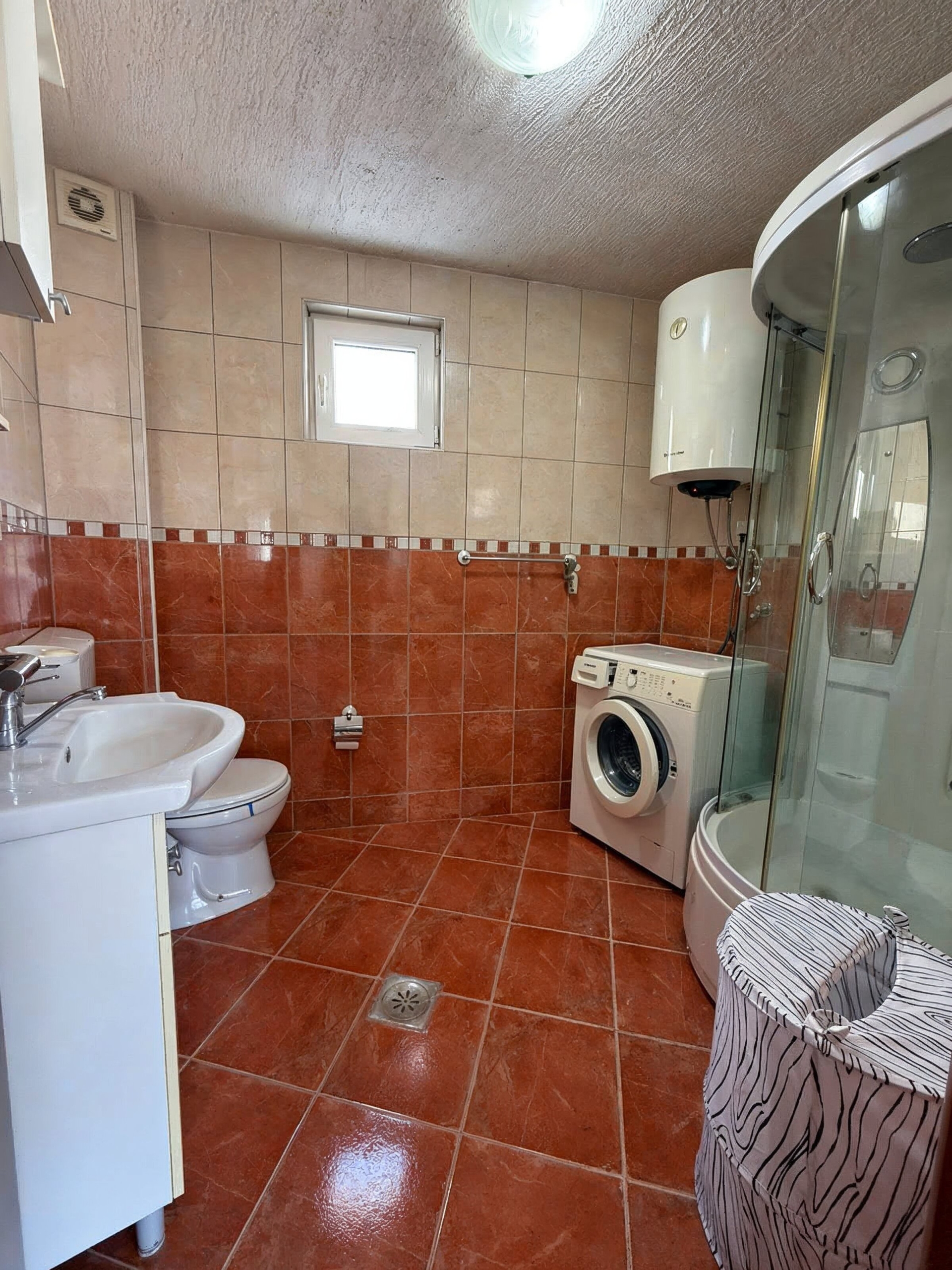 Bathroom in an apartment in Bečići, Montenegro: shower cabin, washing machine, water heater, and terracotta-toned tiles.