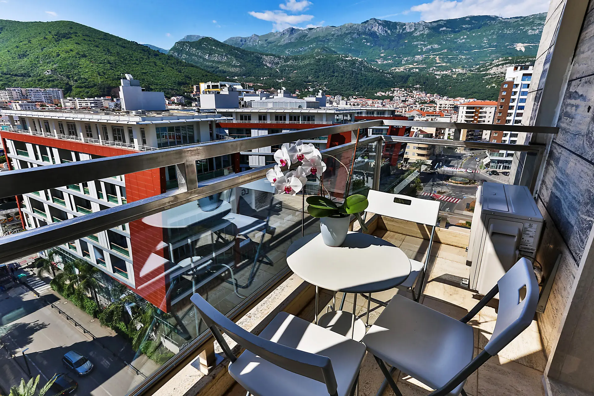 Balcony with furniture and views of the mountains and the city of Budva, Montenegro. On the terrace, a table with an orchid and modern chairs.