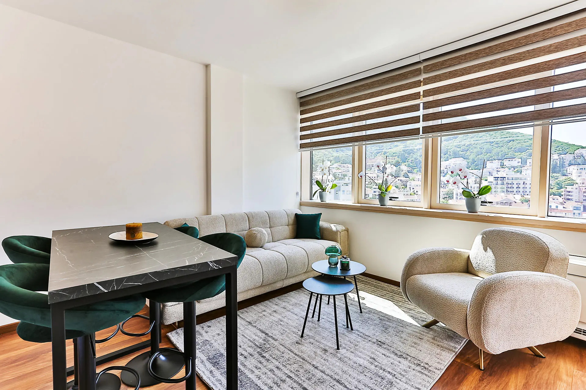 Modern living room in an apartment in Budva, Montenegro: dining area, light-colored sofa, armchair, and mountain view from the window.