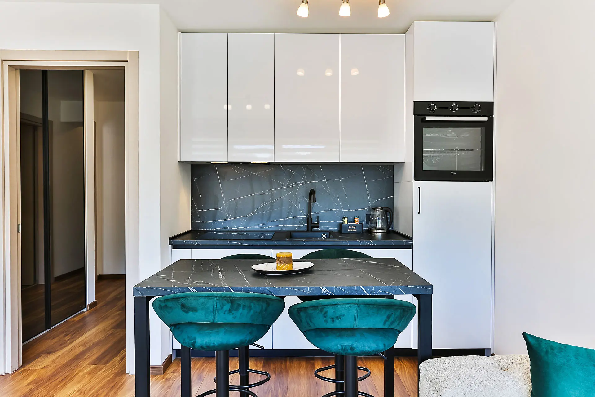 Modern kitchen in an apartment in Budva, Montenegro: white cabinets, a dark marble-look countertop, and emerald bar stools.